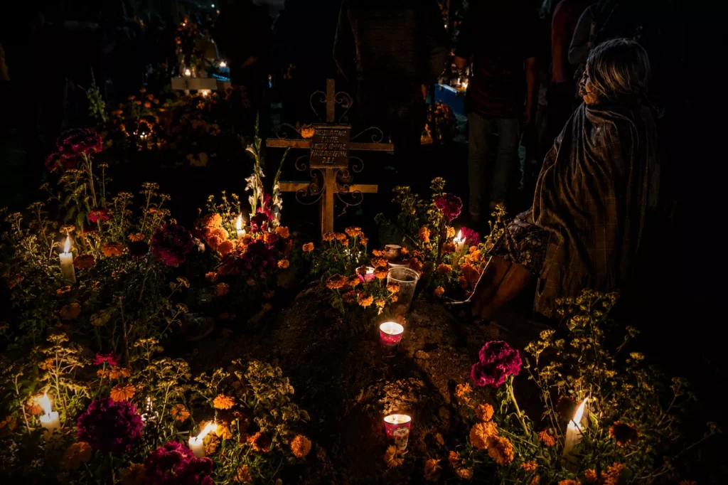 Woman kneels beside candlelit grave covered in flowers